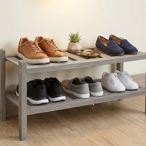 Puede incluir: Un zapatero de madera gris de dos niveles que muestra varios pares de zapatos. El estante superior tiene zapatillas de cuero marr&oacute;n, mocasines azules y una peque&ntilde;a planta. El estante inferior tiene zapatillas negras, grises y blancas.