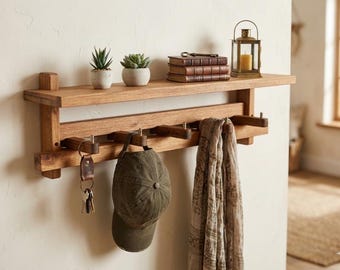 Puede incluir: Una estantería de pared de madera con ganchos, que sostiene una gorra verde, llaves y una bufanda. La estantería también exhibe pequeñas plantas en macetas, libros y una linterna con una vela encendida. La madera tiene un acabado cálido y natural.