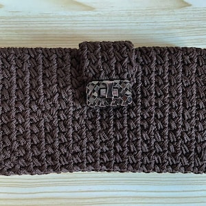 Könnte beinhalten: Eine braune, gewebte Clutch mit rechteckiger Form. Die Clutch hat ein strukturiertes, geflochtenes Muster und einen dekorativen Verschluss in der Mitte. Der Verschluss hat eine dunkle, rechteckige Form mit einem mosaikartigen Design. Die Clutch ist aus weichem Material.