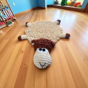May include: A handmade, beige sheep rug with a fluffy texture. The rug features a sheep's head with large, round eyes and a brown, textured woolly head. The sheep's body is a light beige color, and the feet are brown.