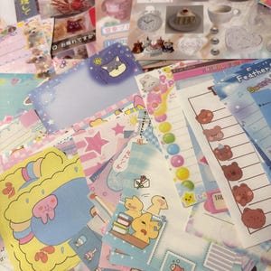May include: A selection of colourful stationery items, including stickers, notepads, and decorative papers. The designs feature cute illustrations of animals, cakes, and stars, with pastel colour schemes. Some items have Japanese text.