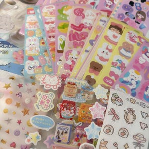 May include: A selection of sticker sheets with diverse designs. The stickers showcase cartoon characters, food, stars, and text. The color scheme is mainly pastel, with pink, blue, yellow, and white. Some stickers have a glittery finish, adding a touch of sparkle.