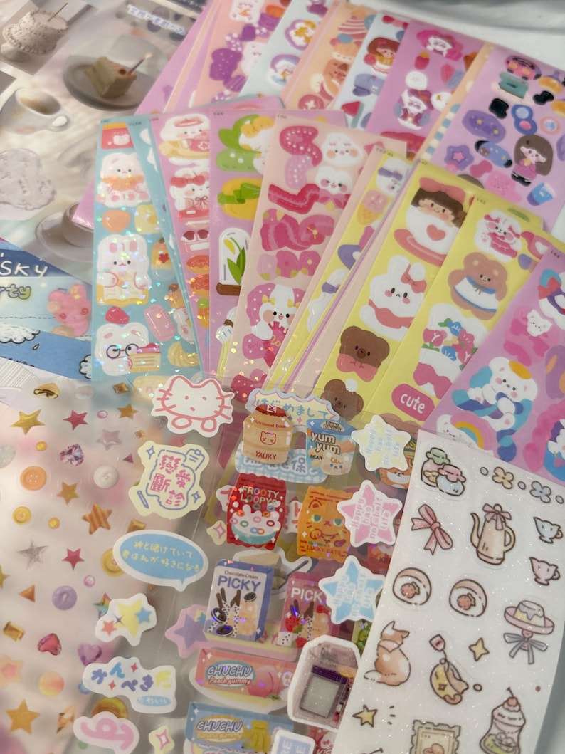 May include: A selection of sticker sheets with diverse designs. The stickers showcase cartoon characters, food, stars, and text. The color scheme is mainly pastel, with pink, blue, yellow, and white. Some stickers have a glittery finish, adding a touch of sparkle.