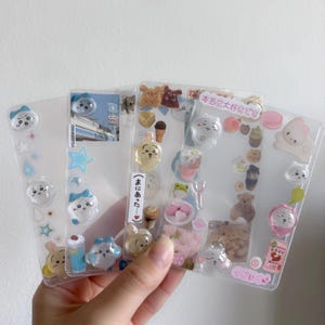 May include: Three clear plastic phone cases decorated with various kawaii-style charms. The charms include cartoon animals, food items like ice cream and macarons, and Japanese text. The cases are held in a hand.