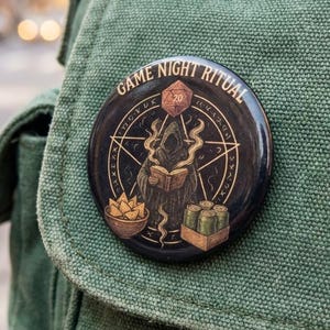 May include: A circular black button with the words "GAME NIGHT RITUAL" around the edge. The button shows a hooded figure reading a book, a bowl of chips, and a box of items. A red 20-sided die is at the top.