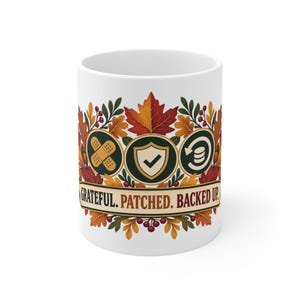 May include: A white ceramic mug featuring a colourful autumn leaf design with red, orange, and yellow foliage. The graphic includes three circular icons: a bandage, a shield with a checkmark, and a data backup symbol, with the text GRATEFUL. PATCHED. BACKED UP.