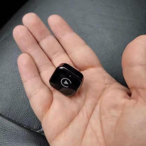 May include: A small, square, black device with rounded corners. A white play symbol is centered on the top surface. The device is held in an open palm.