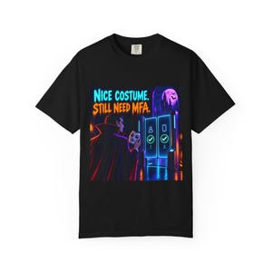 May include: A black t-shirt featuring a graphic of a vampire holding a mask in front of a glowing digital authentication portal. The text reads "NICE COSTUME. STILL NEED MFA." The scene includes a purple moon, bats, and neon blue interface icons.