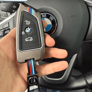 May include: A metallic gray car key with a black strap featuring red, blue, and white stripes. The key has a BMW logo and icons for locking, unlocking, and opening the trunk. The key is held near a black steering wheel.
