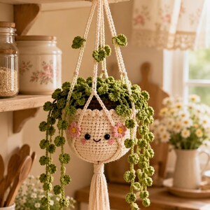 May include: A cream-colored crocheted plant hanger featuring a smiling face with pink cheeks and small pink flowers. The hanger holds a trailing green crocheted plant with round leaves and includes a long, decorative tassel at the bottom.