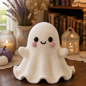 May include: A cream-coloured crochet ghost plush toy featuring a rounded head, two small arms, and a flared base. The ghost has two large black plastic eyes, a small embroidered black mouth, and two circular pink stitched cheeks on its face.
