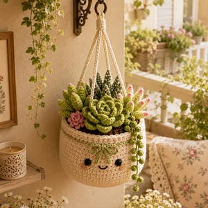 May include: A cream-colored crocheted hanging planter featuring a smiling face with black eyes and pink cheeks. The planter holds a collection of various crocheted succulents in shades of green and pink, suspended by cream-colored yarn cords.