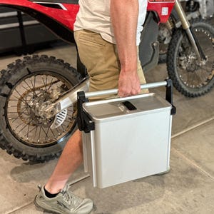 May include: A silver metal container with a black handle is being carried. The container has a rectangular shape and a slot on the top. A red and white dirt bike is in the background.