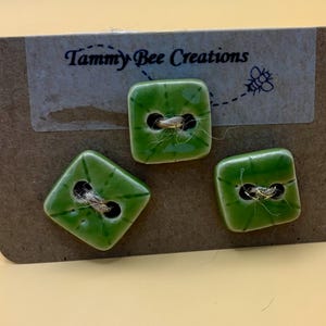 Könnte beinhalten: Drei grüne Keramikknöpfe mit goldenem Faden, präsentiert auf einer braunen Karte. Die Knöpfe sind quadratisch und rautenförmig. Die Karte trägt den Text "Tammy Bee Creations" und eine Bienenillustration.