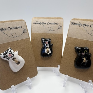 May include: Three cat-shaped buttons on display cards. One button is white with black stripes, another is black with a white chest, and the third is black with a white chest. Each card has the text "Tammy Bee Creations".