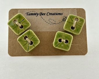 Puede incluir: Cuatro botones cuadrados de cerámica verde claro con líneas grabadas en verde oscuro y dos agujeros, cada uno enhebrado con cordel natural. Los botones están sujetos a una tarjeta de presentación rectangular marrón con el texto Tammy Bee Creations.