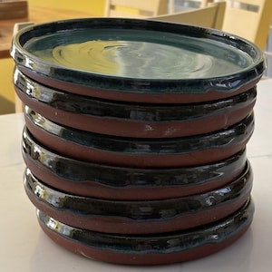 Puede incluir: Una pila de cinco platos redondos de cerámica con una base de arcilla de color marrón rojizo oscuro. Cada plato tiene un esmalte brillante de color verde azulado profundo y negro en la superficie interior y el borde exterior, con acabado liso.