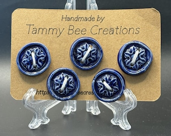 Puede incluir: Cinco botones de cerámica azul oscuro de forma redonda con un patrón floral en relieve en el centro y dos agujeros para el hilo. Los botones están sujetos a una tarjeta rectangular marrón con la etiqueta "Handmade by Tammy Bee Creations".