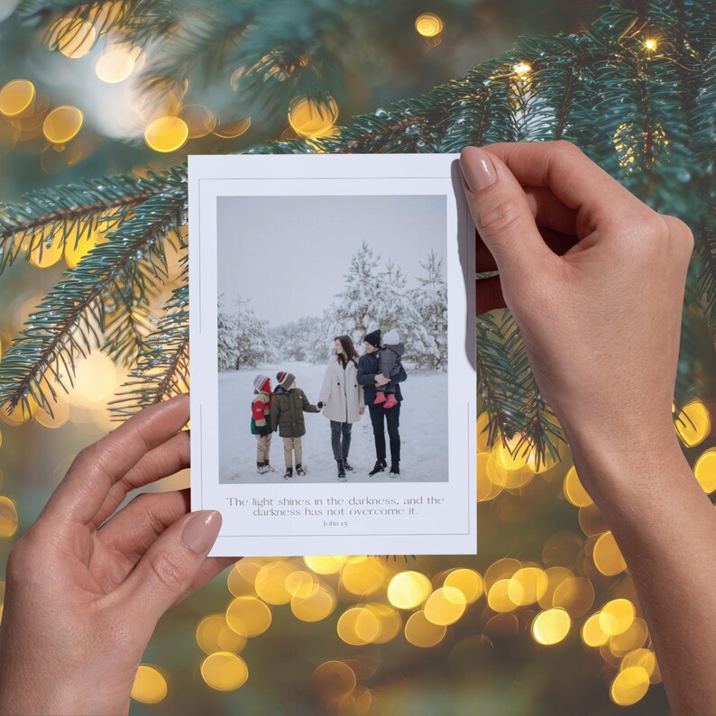 May include: A white Christmas card held by hands, featuring a family portrait in a snowy landscape. The card has a thin border and includes the text: "The light shines in the darkness, and the darkness has not overcome it."