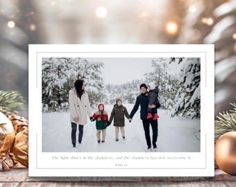 May include: A family portrait on a white card with a thin gray border. The image shows a family of five walking in a snowy forest. The text reads, "The light shines in the darkness, and the darkness has not overcome it. John 1:5."