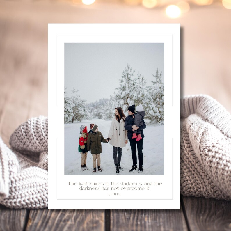 May include: A family of five stands in a snowy forest, the parents holding hands with their children. The image is framed by a white border and includes the text "The light shines in the darkness, and the darkness has not overcome it. John 1:5."