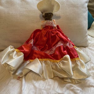 May include: A doll in a red satin dress with white lace trim and a white chef's hat. The dress features white satin sleeves and gold trim. The doll is positioned on a white surface.