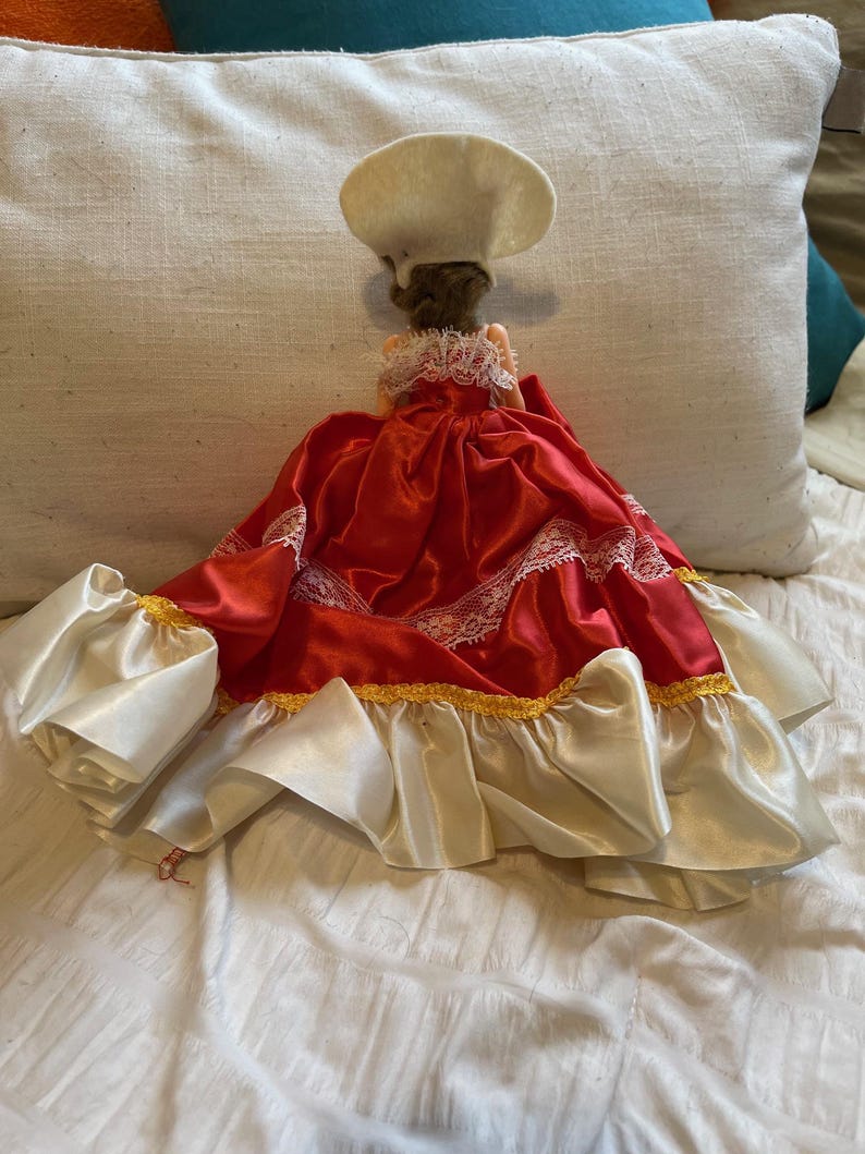 May include: A doll in a red satin dress with white lace trim and a white chef's hat. The dress features white satin sleeves and gold trim. The doll is positioned on a white surface.