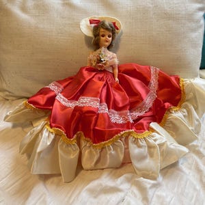 May include: A vintage doll in a red satin dress with white lace trim and a cream-coloured skirt. The doll wears a matching hat with a red bow and holds a small bouquet. A collectible item.