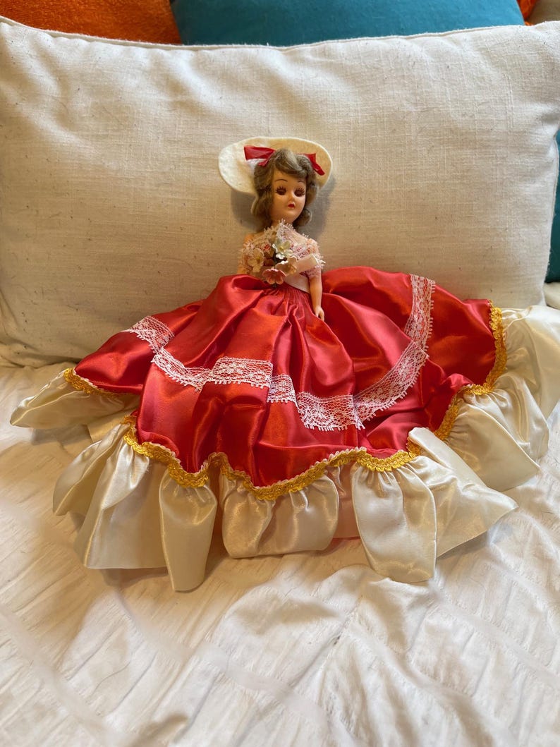 May include: A vintage doll in a red satin dress with white lace trim and a cream-coloured skirt. The doll wears a matching hat with a red bow and holds a small bouquet. A collectible item.