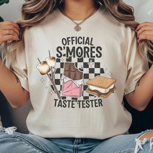 May include: Cream-colored t-shirt with the text "OFFICIAL S'MORES TASTE TESTER" and a graphic of a s'more, chocolate bar, and marshmallows on sticks. The design includes a black and white checkered background.