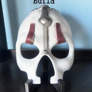 May include: A white skull-shaped mask featuring a central silver vertical ridge with a circular accent, two dark red rectangular markings above the eye sockets, and the text "Quick one-day build" displayed above the object.