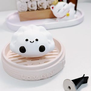 Peut inclure: Un diffuseur d'air en forme de ravioli blanc avec un visage souriant, posé sur une base rose en forme de cuiseur vapeur. Une petite figurine de chat blanc avec une couronne dorée et une bouteille d'huile essentielle brune sont en arrière-plan.