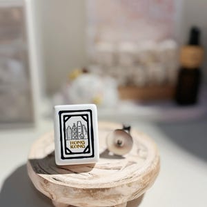 May include: A white rectangular tile with a black border and the words "HONG KONG" in gold lettering. The tile features a black and white illustration of Hong Kong's skyline. The tile is on a small, round, wooden tray.