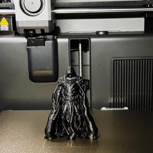 Puede incluir: Una figura negra impresa en 3D de un guerrero con capa, de pie sobre una superficie texturizada dentro de una impresora 3D. La figura tiene detalles intrincados y es el punto focal. La impresora es negra y gris.