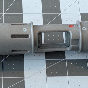 May include: A gray lightsaber hilt with black ends and a rectangular opening in the center. The hilt has a textured surface and a small red accent. The lightsaber is lying on a gray and black grid.