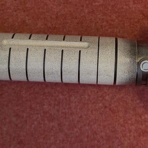 Puede incluir: Un accesorio de sable de luz detallado con un acabado met&aacute;lico. El mango presenta una combinaci&oacute;n de tonos negros y grises, con una secci&oacute;n central en blanco con l&iacute;neas verticales negras. La hoja emite un brillo rojo brillante.