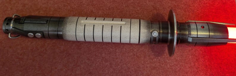 Puede incluir: Un accesorio de sable de luz detallado con un acabado met&aacute;lico. El mango presenta una combinaci&oacute;n de tonos negros y grises, con una secci&oacute;n central en blanco con l&iacute;neas verticales negras. La hoja emite un brillo rojo brillante.