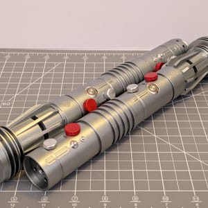 Puede incluir: Dos sables de luz plateados con botones rojos y plateados. Los sables de luz tienen un dise&ntilde;o cil&iacute;ndrico con crestas detalladas y un extremo circular. Los sables de luz est&aacute;n sobre una esterilla de corte gris.