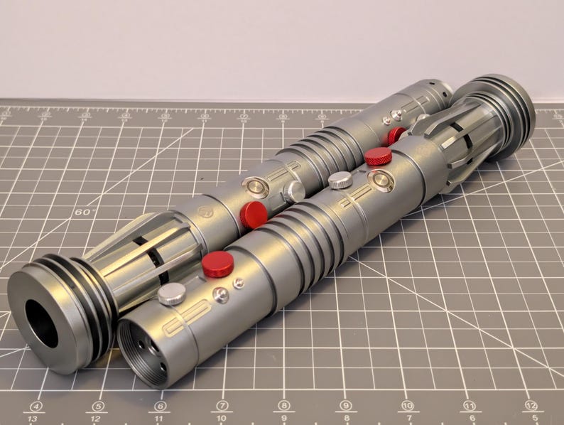 Puede incluir: Dos sables de luz plateados con botones rojos y plateados. Los sables de luz tienen un dise&ntilde;o cil&iacute;ndrico con crestas detalladas y un extremo circular. Los sables de luz est&aacute;n sobre una esterilla de corte gris.