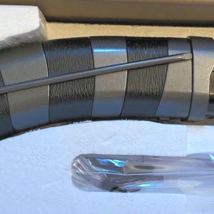 May include: A silver and black lightsaber hilt with a curved handle and a purple blade. The handle features a textured grip and a series of silver and black bands. The lightsaber is packaged in a cardboard box with white foam inserts.