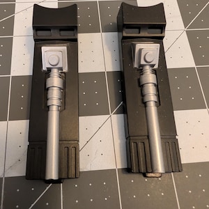 May include: Two black, rectangular components with silver-colored cylindrical and square details. The components have a stepped design and are placed on a gray grid surface. The silver parts have a button-like feature.