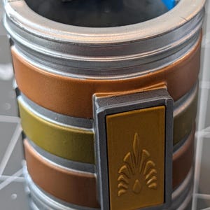 May include: A cylindrical object with silver, bronze, and gold bands. A rectangular gold emblem with a stylized leaf design is on the side. The object has a metallic finish and a hollow interior.