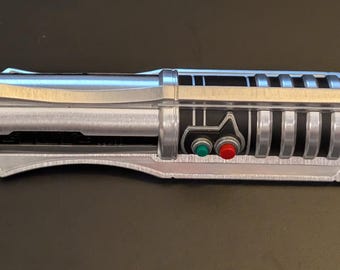 May include: A silver and black lightsaber prop with a detailed design. The hilt features a silver finish with black accents, including a control panel with red and green buttons. The blade is silver and flat.