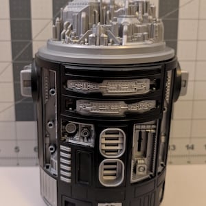May include: A detailed, cylindrical, black and silver model with intricate mechanical detailing. The top features a silver cityscape design. The model has a futuristic aesthetic, with various panels and vents.