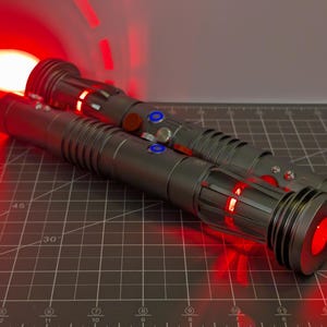 Puede incluir: Dos sables de luz grises con detalles rojos y azules, uno con una hoja roja brillante. Los mangos tienen dise&ntilde;os detallados con botones y luces. El fondo es una esterilla de corte oscura.