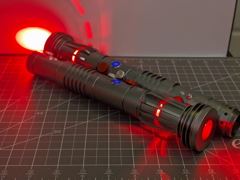 Puede incluir: Dos sables de luz grises con detalles rojos y azules, uno con una hoja roja brillante. Los mangos tienen dise&ntilde;os detallados con botones y luces. El fondo es una esterilla de corte oscura.