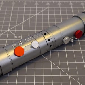 Puede incluir: Un accesorio de sable de luz plateado con botones rojos y plateados. El sable de luz tiene un dise&ntilde;o cil&iacute;ndrico con una serie de crestas y agujeros a lo largo de su cuerpo. Los botones son rojos y plateados.