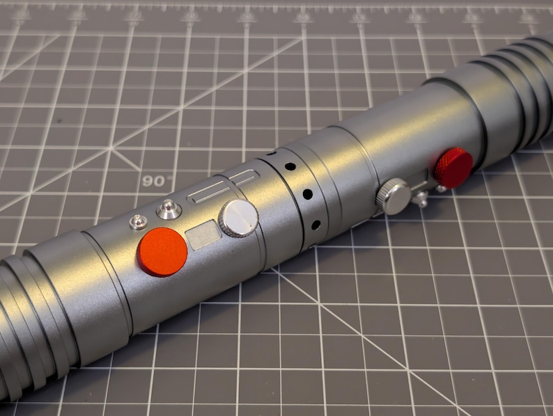 Puede incluir: Un accesorio de sable de luz plateado con botones rojos y plateados. El sable de luz tiene un dise&ntilde;o cil&iacute;ndrico con una serie de crestas y agujeros a lo largo de su cuerpo. Los botones son rojos y plateados.