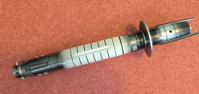 Puede incluir: Un accesorio de sable de luz met&aacute;lico con un mango blanco y negro y un protector de hoja plateado. El sable de luz tiene un dise&ntilde;o detallado con un cuerpo cil&iacute;ndrico y un emisor acampanado.