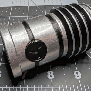 May include: A silver and black lightsaber hilt. The hilt has a cylindrical design with ribbed black sections and a black button. The end of the hilt is a flat, rectangular shape.